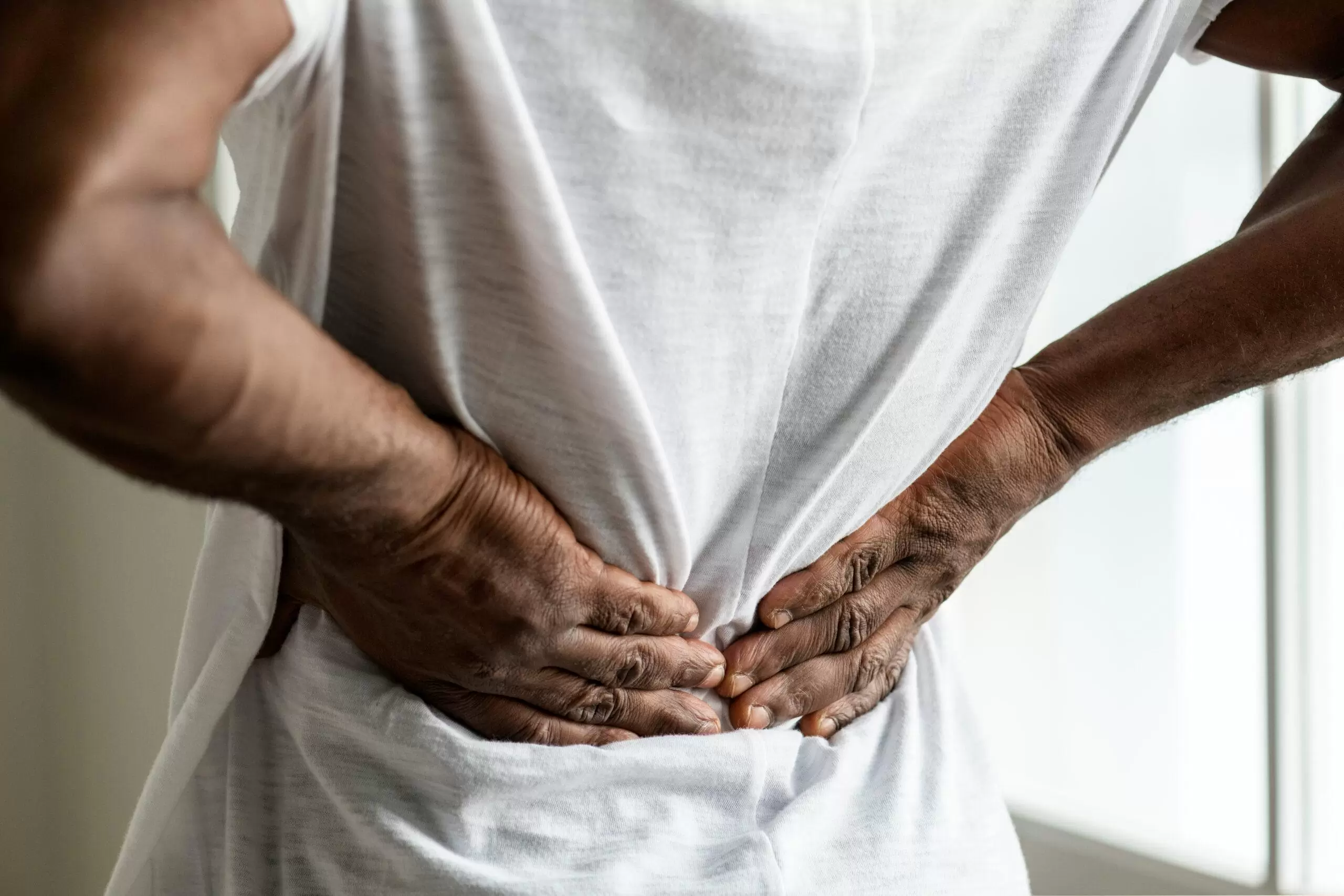 Close-up of a person gripping their lower back with both hands, appearing to be in pain or experiencing back stiffness